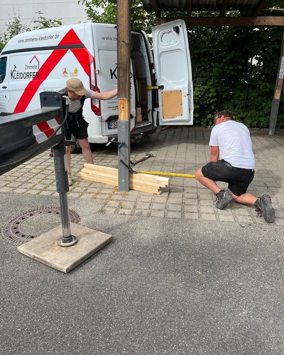 Zwei Handwerker bei der Arbeit, einer misst Holzbretter, der andere lehnt sich an einen Holzpfosten.