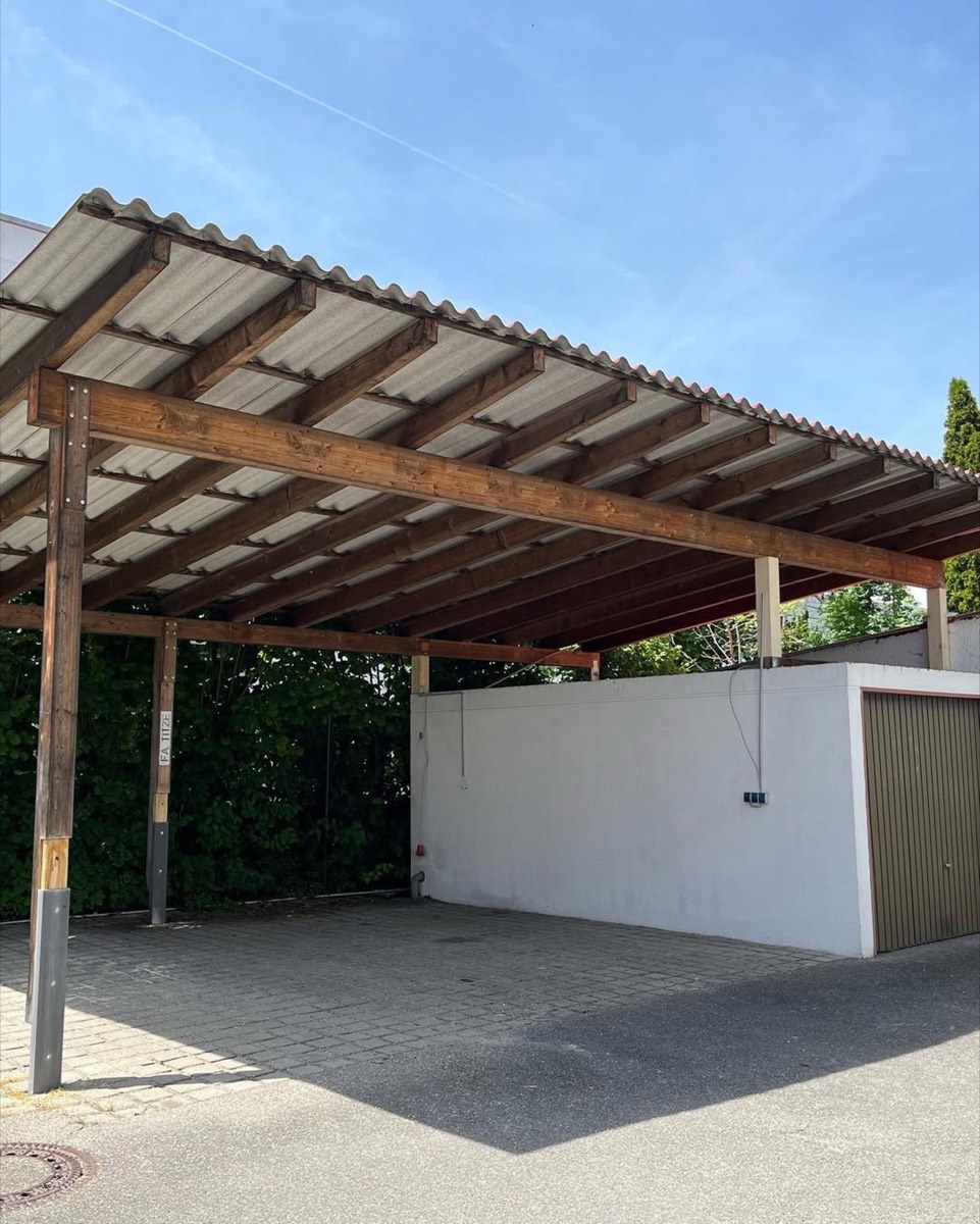 Ein Carport aus Holz mit Wellblechdach über einem gepflasterten Parkplatz neben einer weißen Wand und einer Garage.