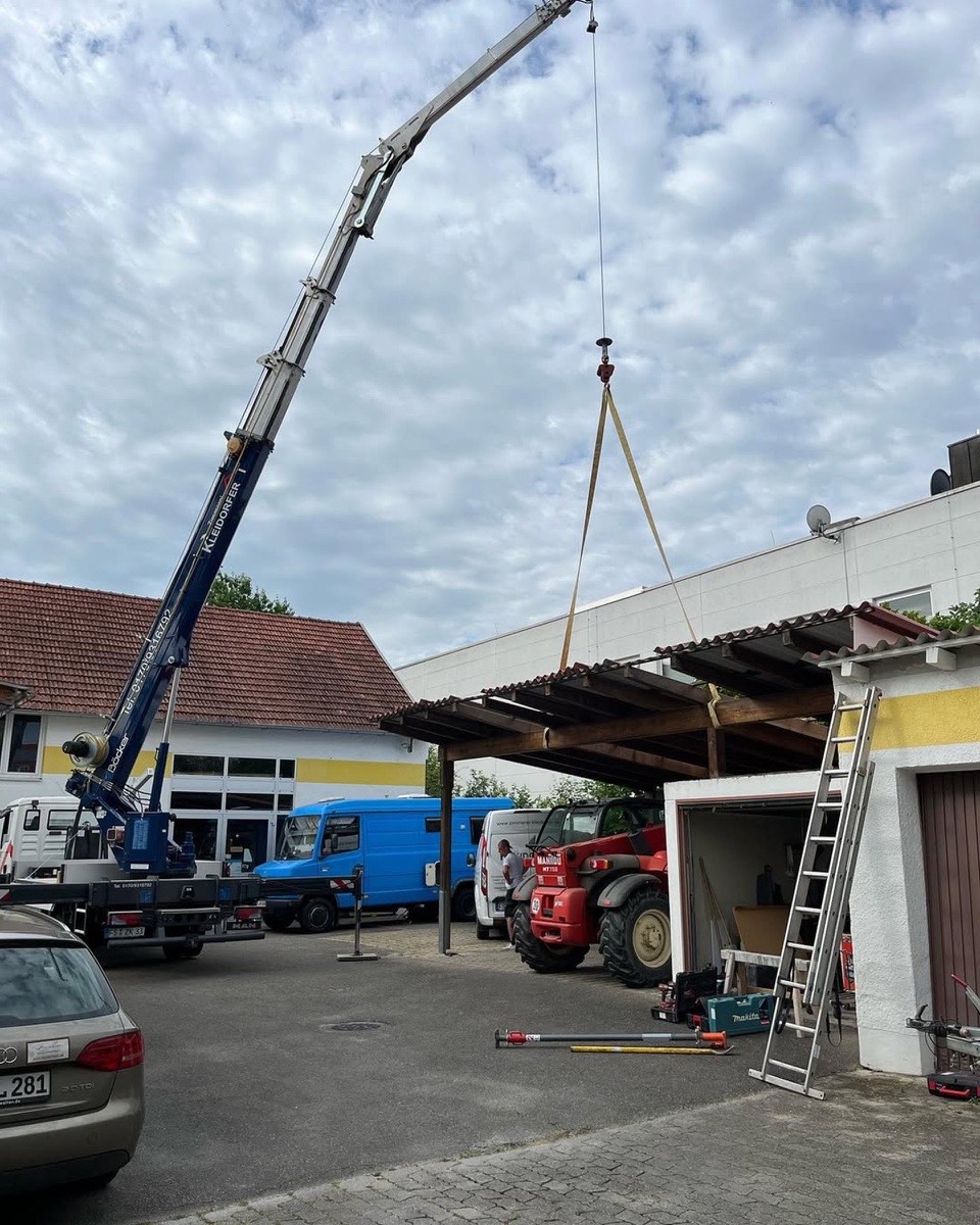 Ein großer blauer Kran hebt ein dunkles Carport-Dach mit Gurten über einem Hof mit Fahrzeugen.