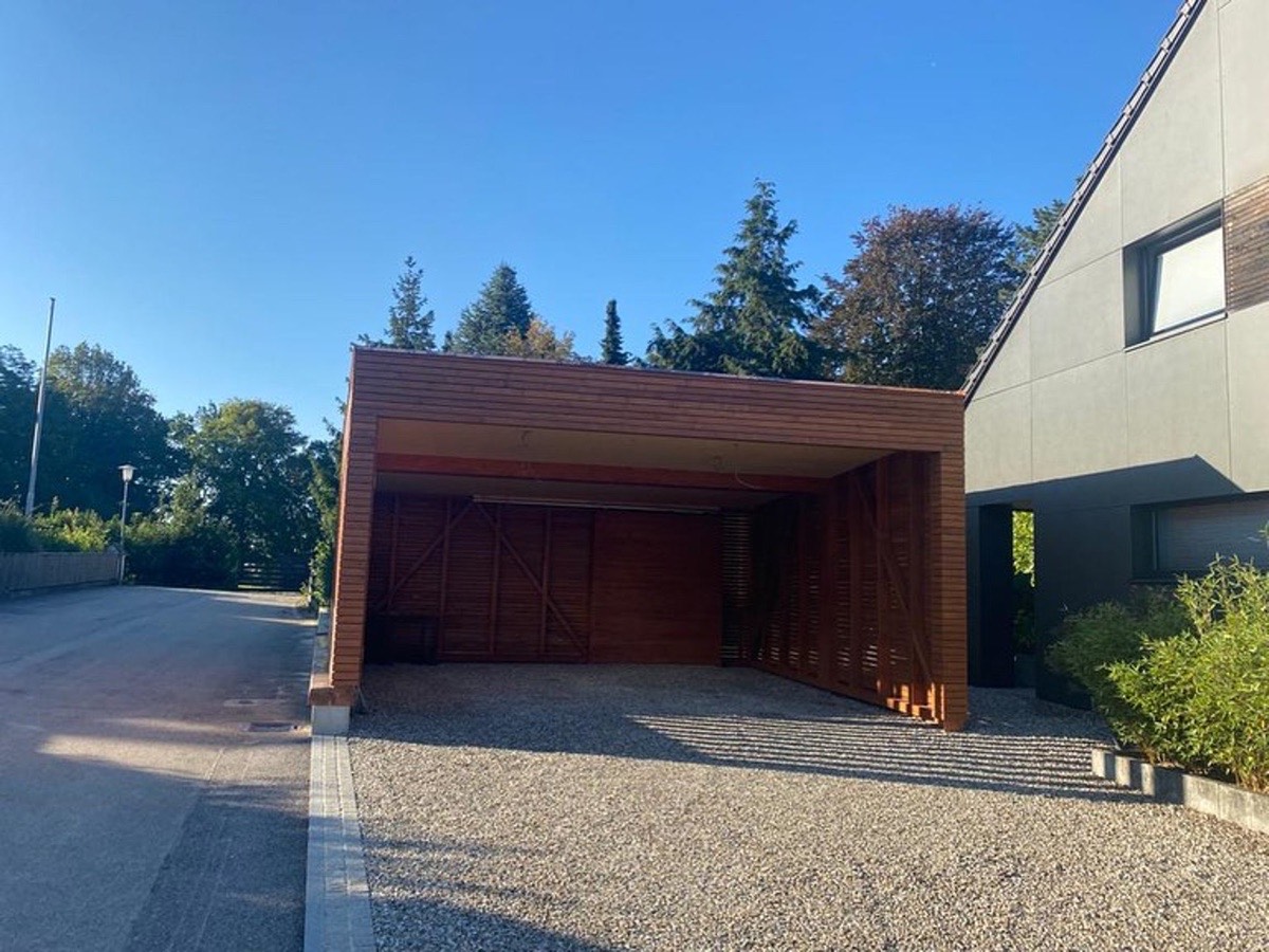 Modernes Carport aus Holz mit Kiesauffahrt neben einem Haus und einer Straße an einem sonnigen Tag.