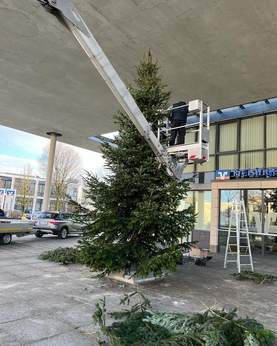 Ein Mann in einem Hebekran schmückt einen großen Weihnachtsbaum vor einem Gebäude mit der Aufschrift „Volksbank“.