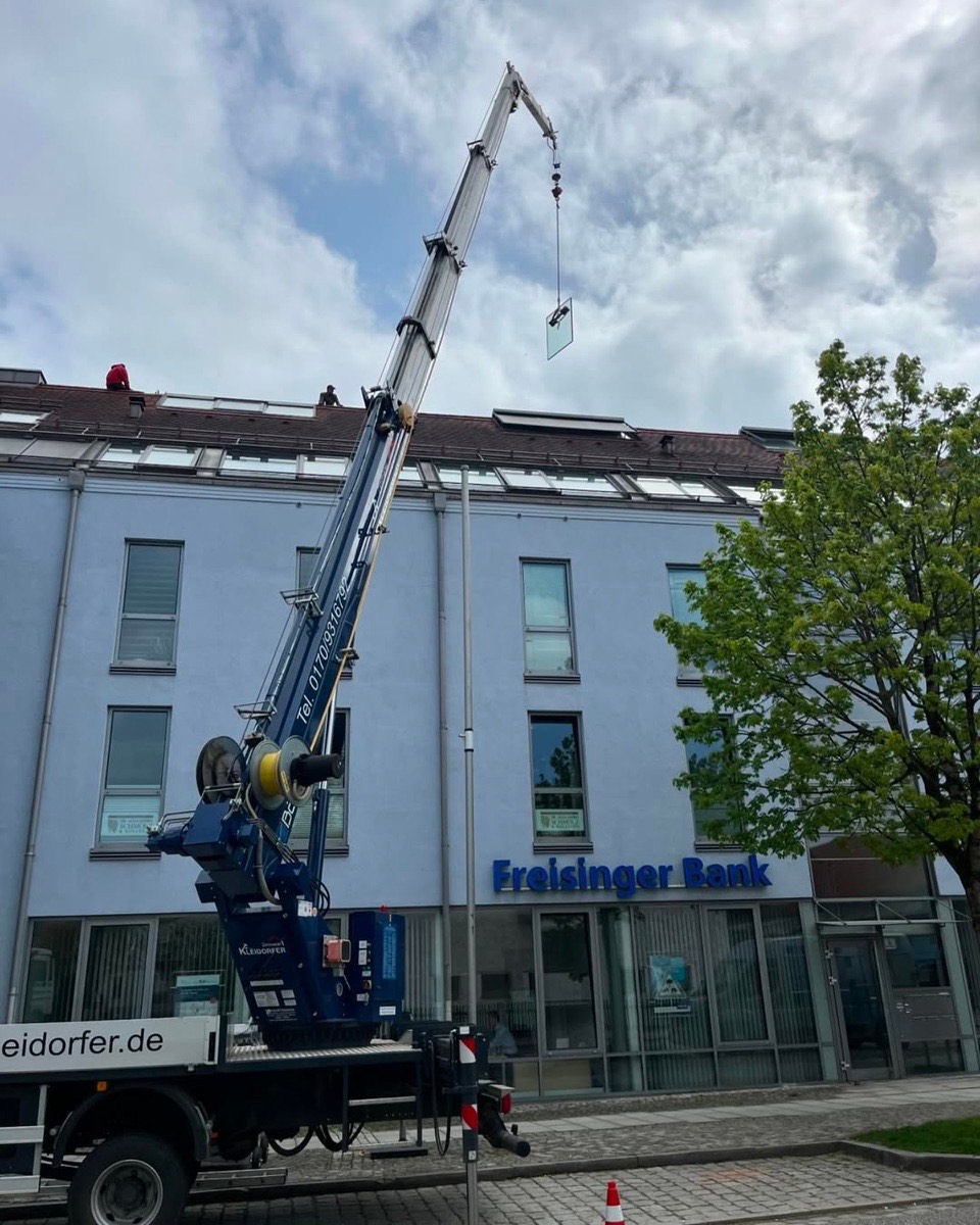 Ein blauer Kranwagen hebt eine Glasscheibe vor einem Gebäude mit der Aufschrift „Freisinger Bank“ auf ein Dach.