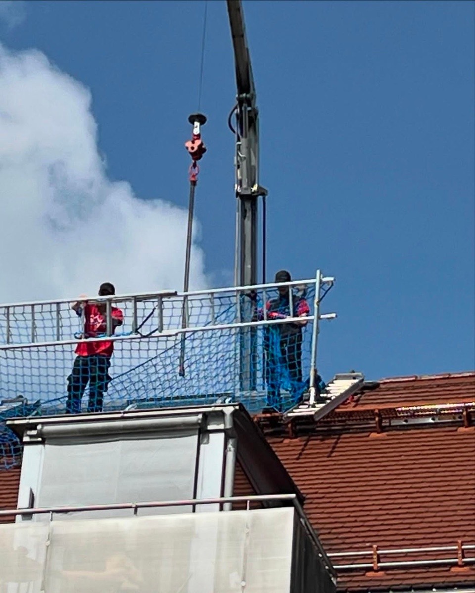 Zwei Bauarbeiter in Schutzkleidung stehen auf einem Gerüst neben einem Kranarm vor einem Ziegeldach.