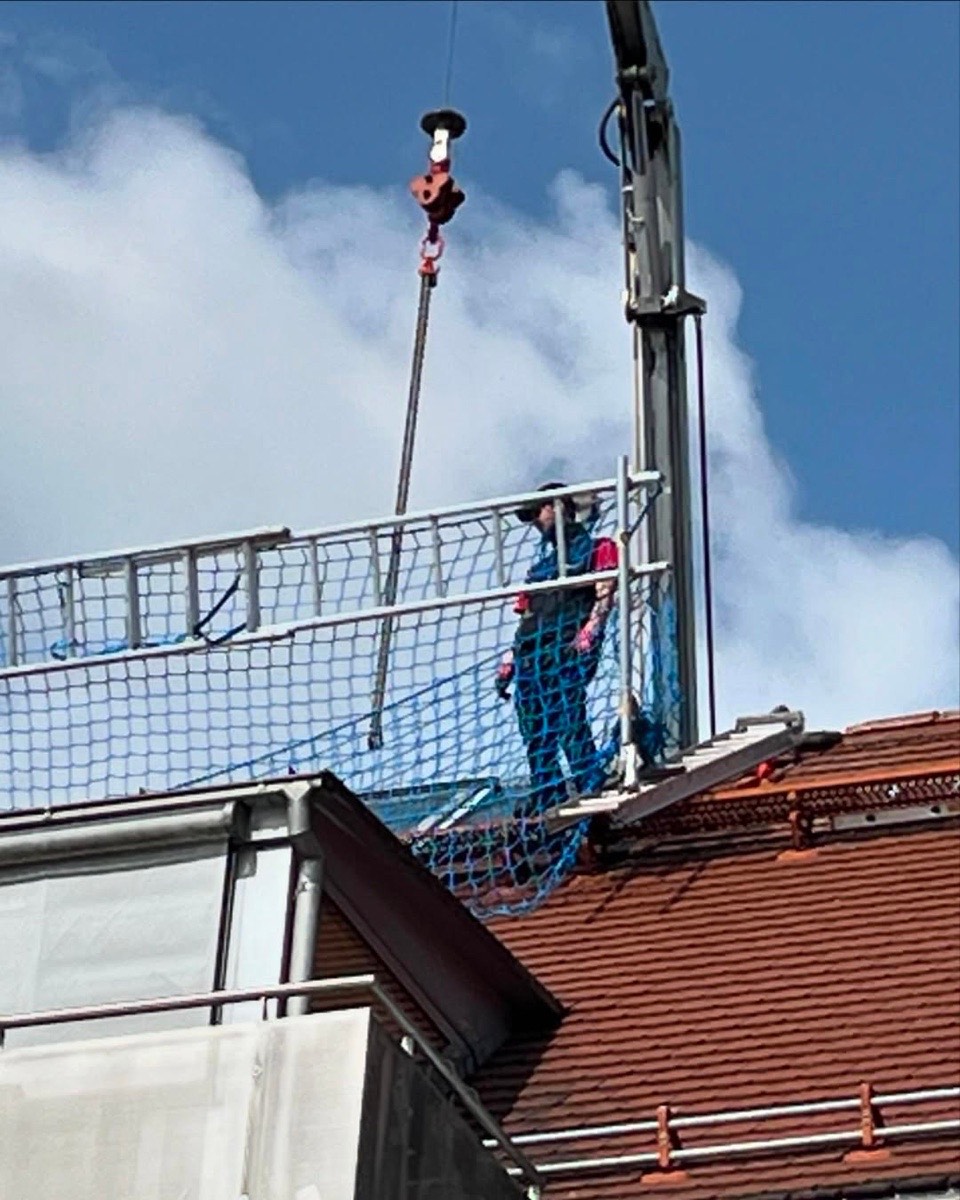 Ein Arbeiter auf einem Gerüst mit blauem Netz steht auf einem Dach und bedient einen Kranhaken unter bewölktem Himmel.