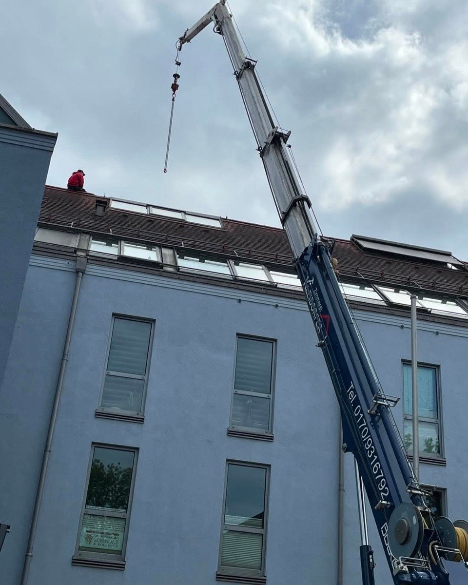 Ein blauer Kran mit einem langen Ausleger ragt über ein Gebäude mit Dachfenstern, eine Person sitzt auf dem Dach.