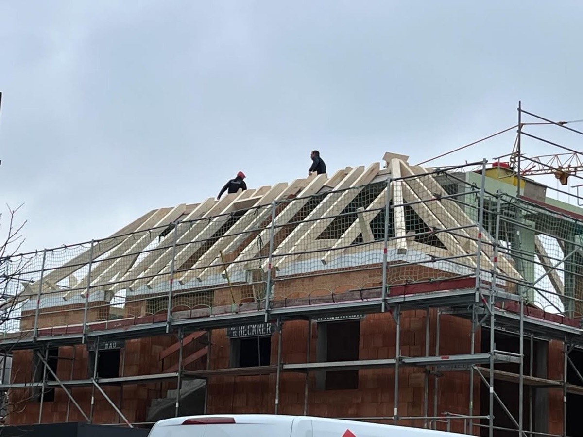 Zwei Dachdecker arbeiten an einem Hausdach mit Holzbalken und Gerüst unter bewölktem Himmel.