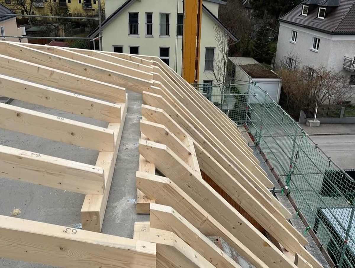 Neues Holzdachstuhl-Gerüst auf einem Rohbau mit Häusern und Gerüstnetz im Hintergrund.