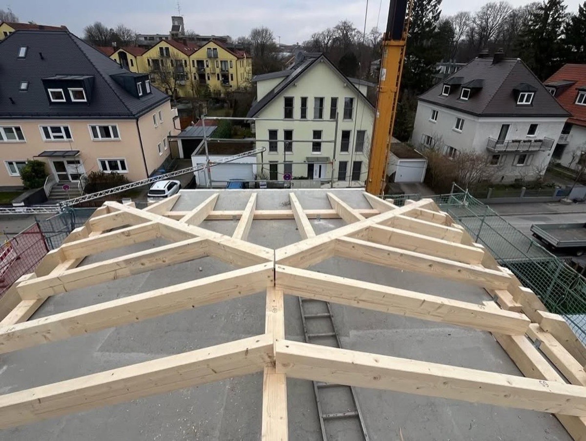 Dachstuhl aus hellem Holz auf einem neuen Gebäude, mit Kran und Häusern im Hintergrund.