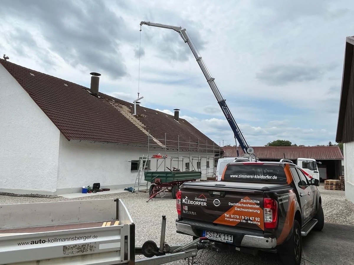 Ein Nissan Pick-up mit Kran und Anhänger vor einem Gebäude, dessen Dach repariert wird.