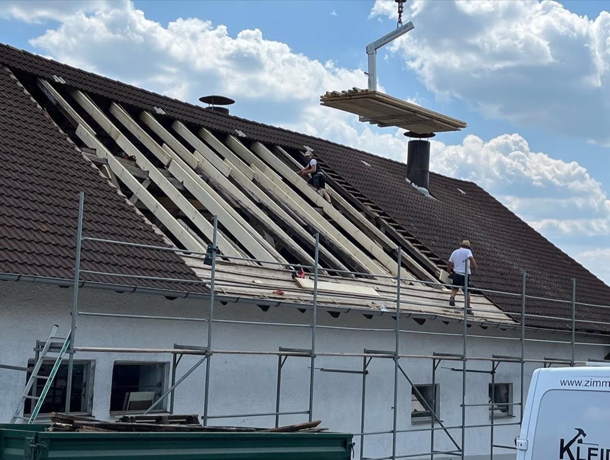 Dachdecker montieren Dachlatten auf einem braunen Ziegeldach, während ein Kran Holzbretter liefert.