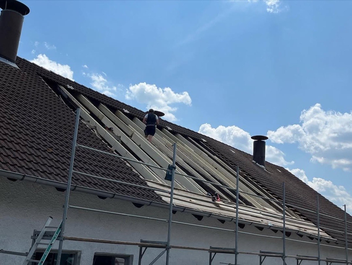 Dachdecker arbeitet auf einem Dach mit freigelegten Holzlatten und entfernt Ziegel, Gerüst darunter.