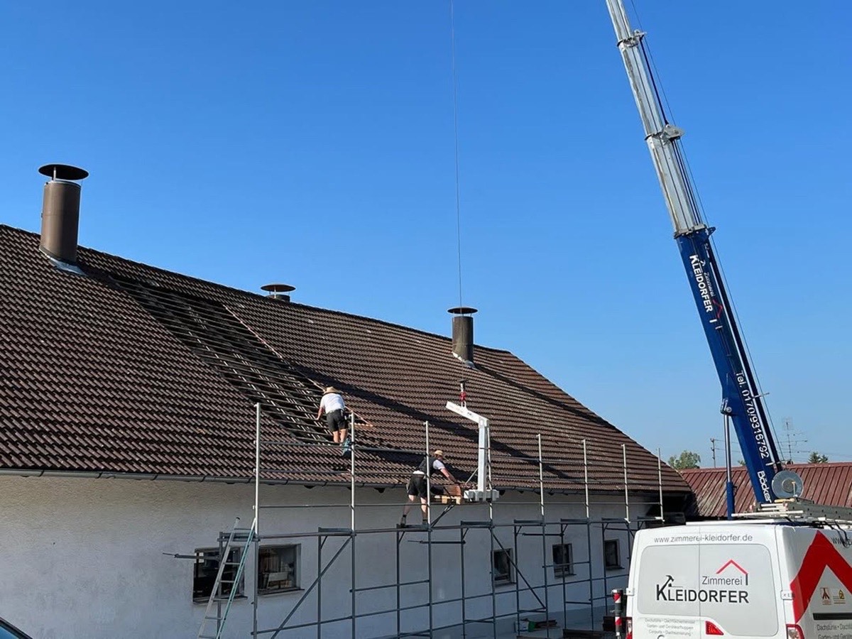 Zwei Dachdecker auf einem Hausdach mit braunen Ziegeln, während ein Kran ein Rohr hebt.