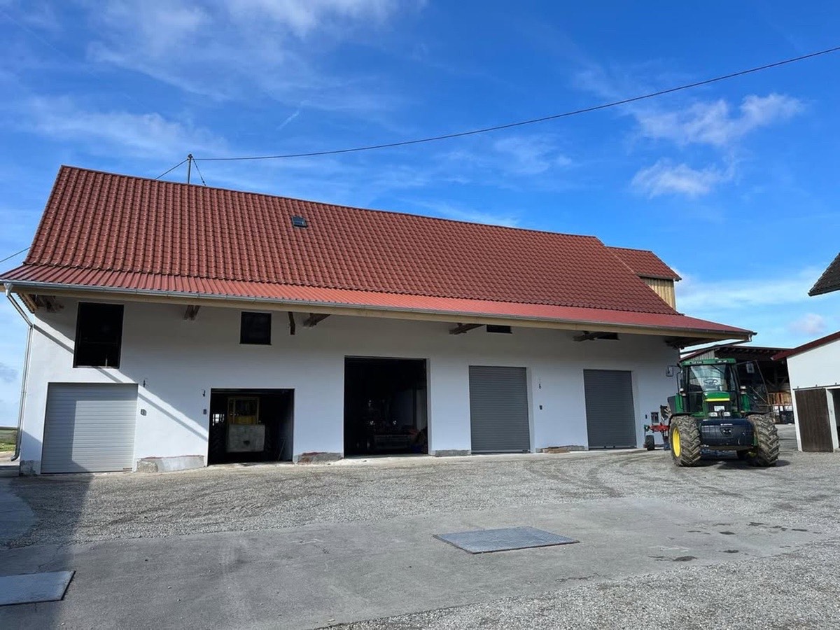 Weißes Bauernhaus mit rotem Ziegeldach, vier dunklen Garagentoren und grünem Traktor davor unter blauem Himmel.