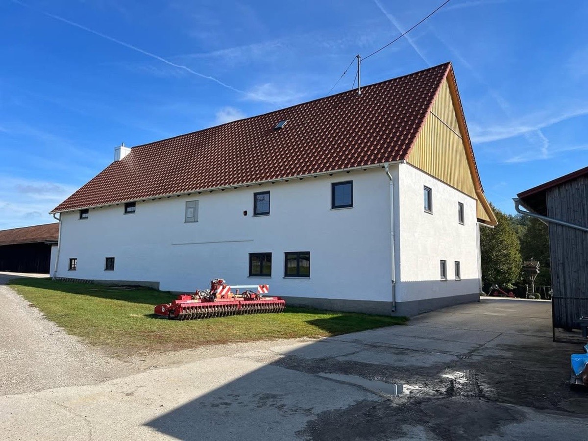Weißes Bauernhaus mit braunem Ziegeldach und einem roten Landwirtschaftsgerät auf dem Rasen.