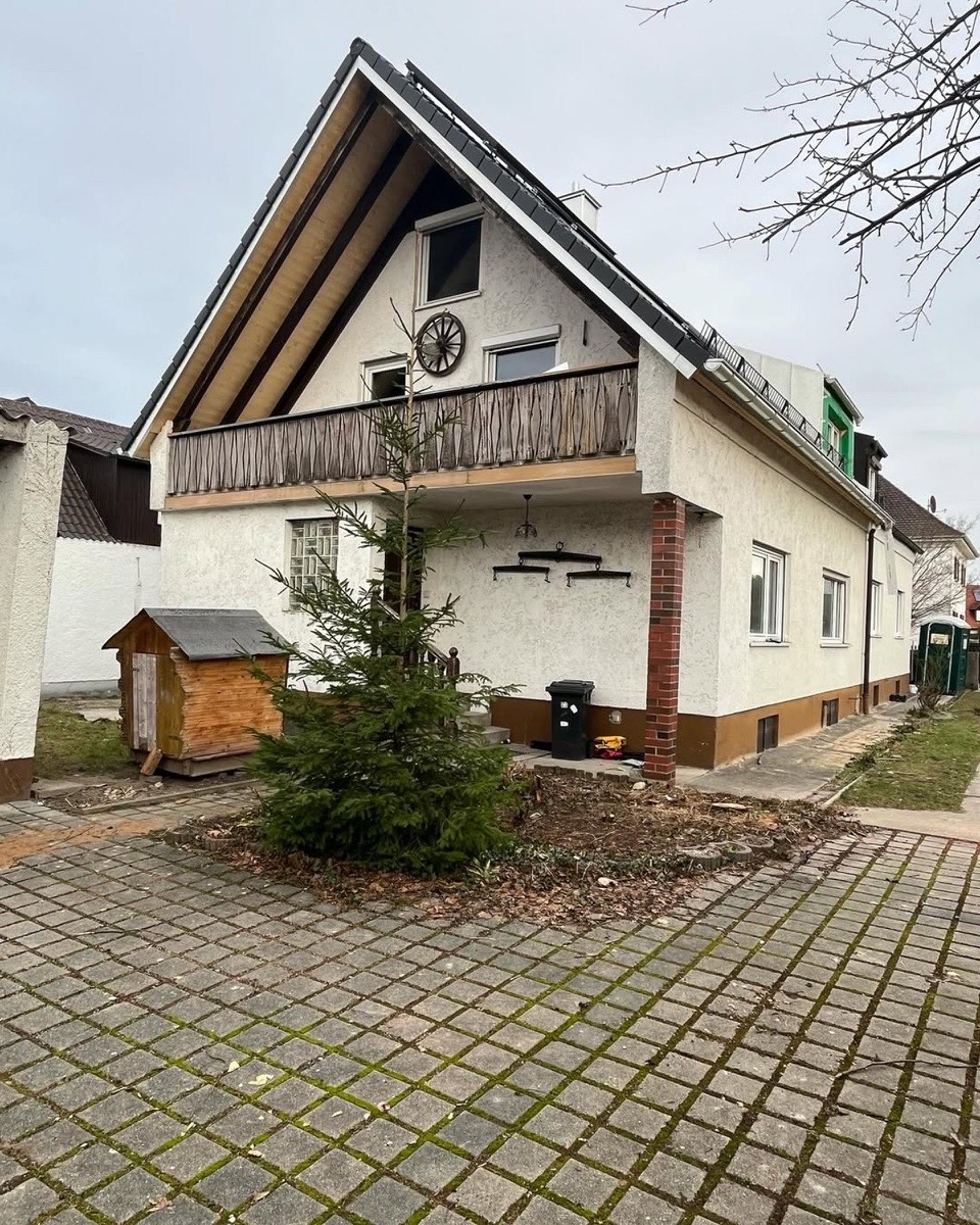 Weißes Haus mit Giebel, Balkon, schwarzem Dach, Weihnachtsbaum, Hundehütte und Pflastersteinen.