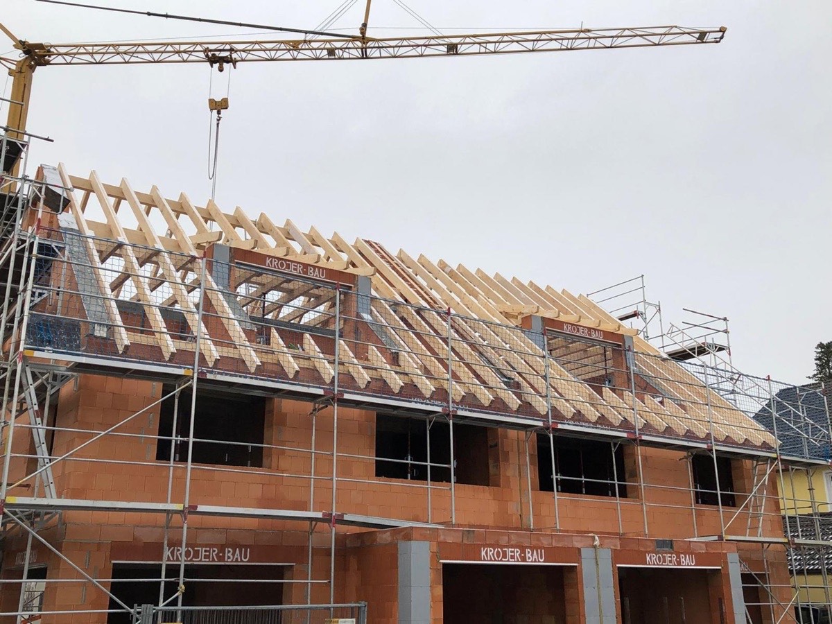 Ein im Bau befindliches Haus mit Dachstuhl, Gerüst und Kran vor grauem Himmel.