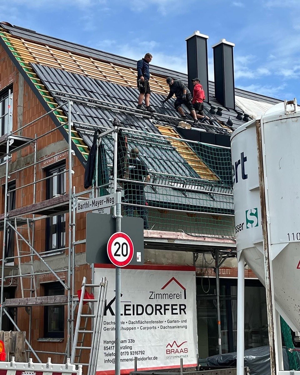 Drei Dachdecker arbeiten an der Eindeckung eines Ziegeldachs mit zwei Schornsteinen; ein 20 km/h Schild und ein Firmenschild sind sichtbar.