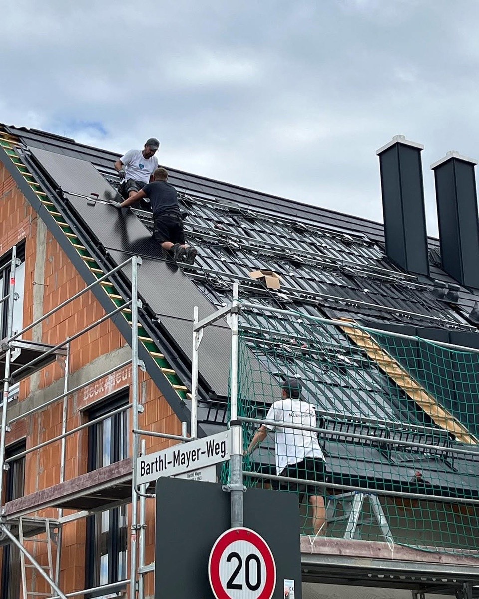 Drei Arbeiter installieren Solarmodule auf einem Ziegeldach. Ein Straßenschild zeigt "Barthl-Mayer-Weg" und ein Tempolimit von 20 an.