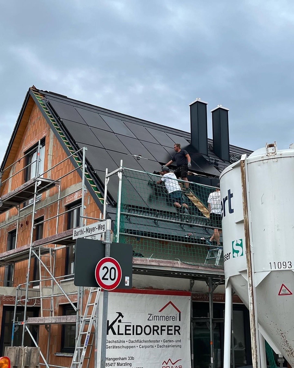 Drei Bauarbeiter installieren Solarpaneele auf dem Dach eines Hauses mit Gerüst und einem Bauschild.