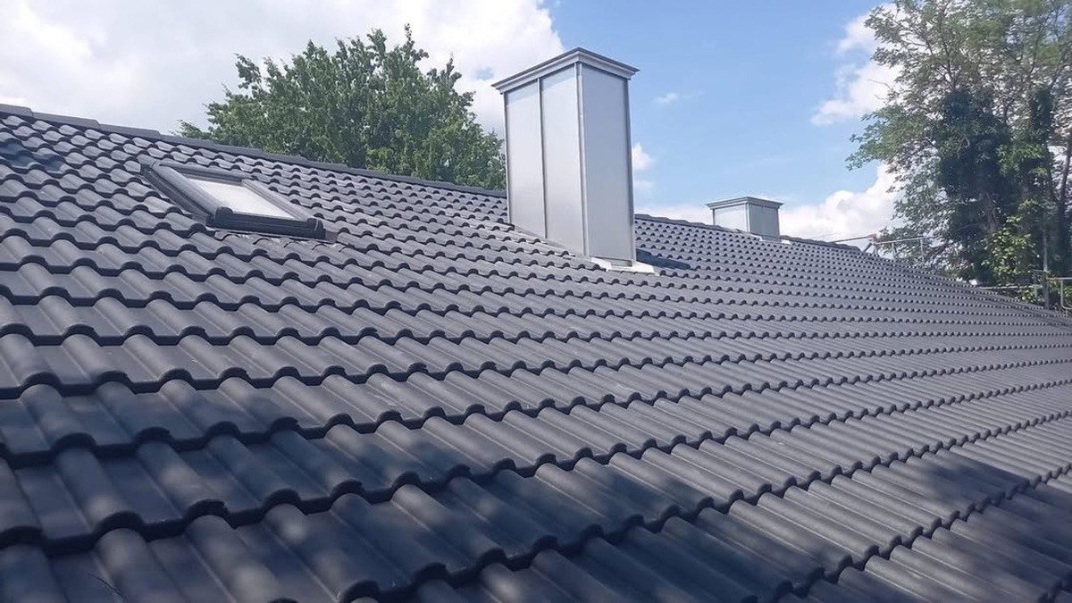 Dunkelgraues Ziegeldach mit einem Dachfenster und zwei silbernen Schornsteinen unter blauem Himmel.